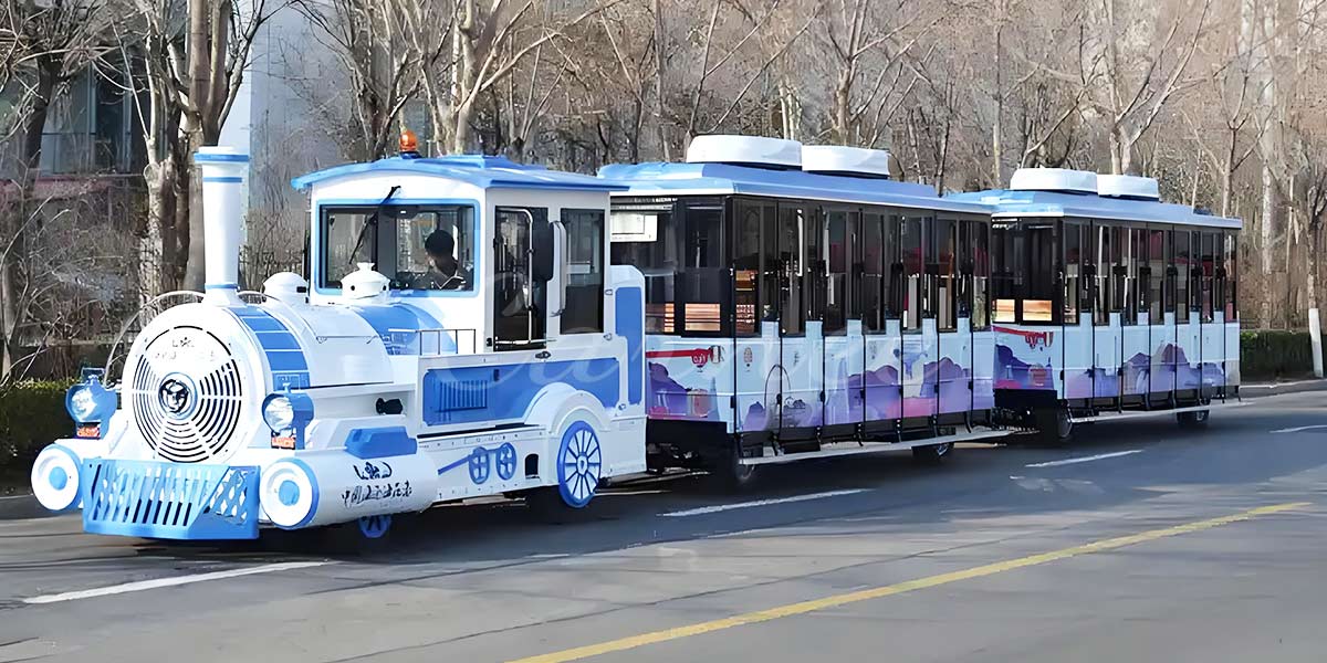 A modern blue and white trackless train carrying passengers on a city street, featuring a stylish locomotive and two enclosed carriages.