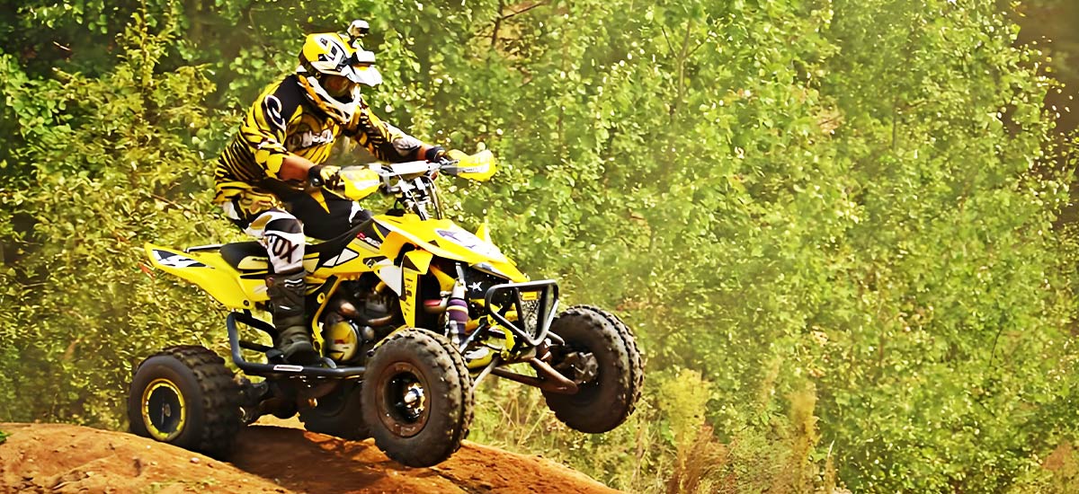 A rider performing a jump on a yellow ATV in a lush forest trail.
